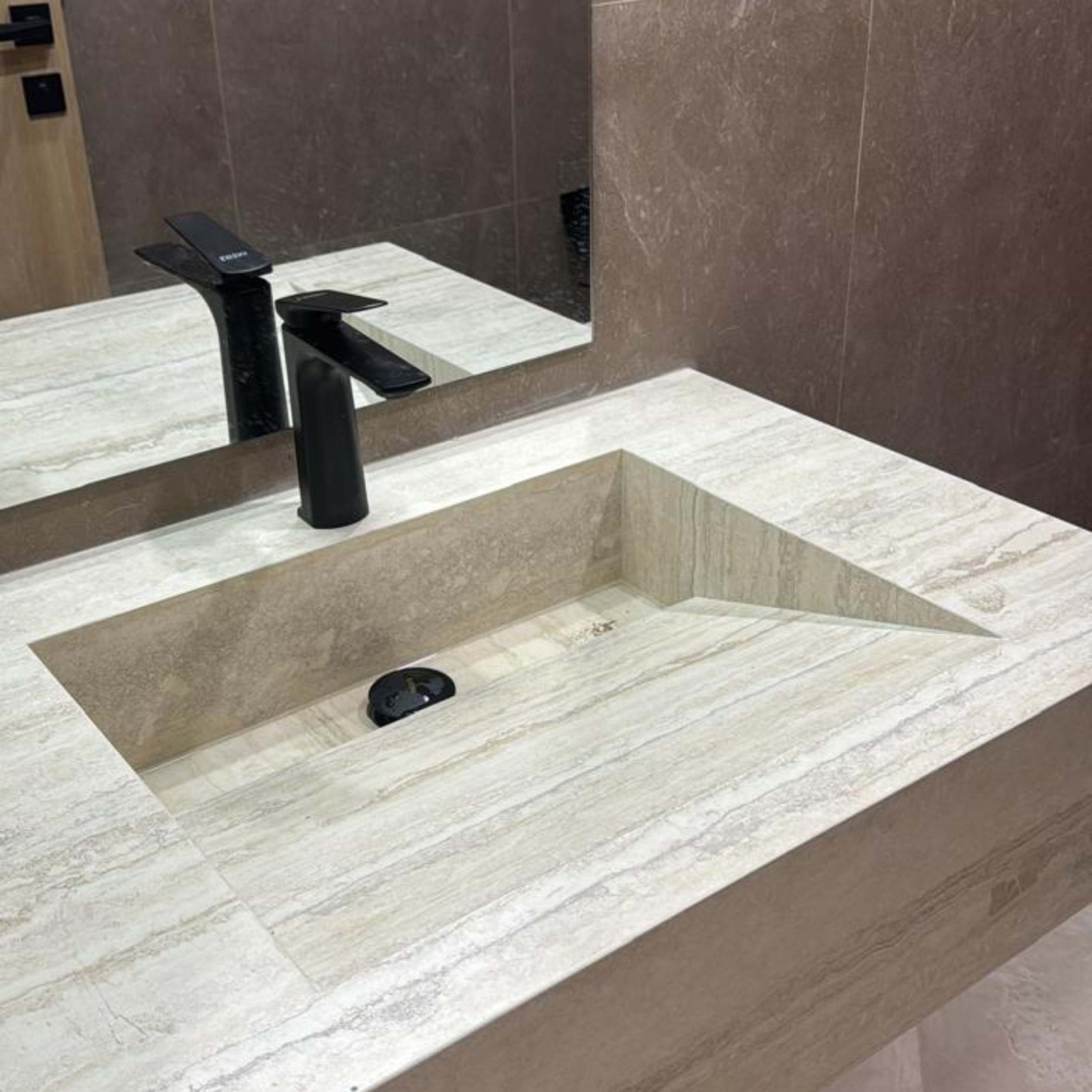 A close-up of an integrated beige travertine stone bathroom sink with a distinct sloped basin design and a sleek matte black faucet.