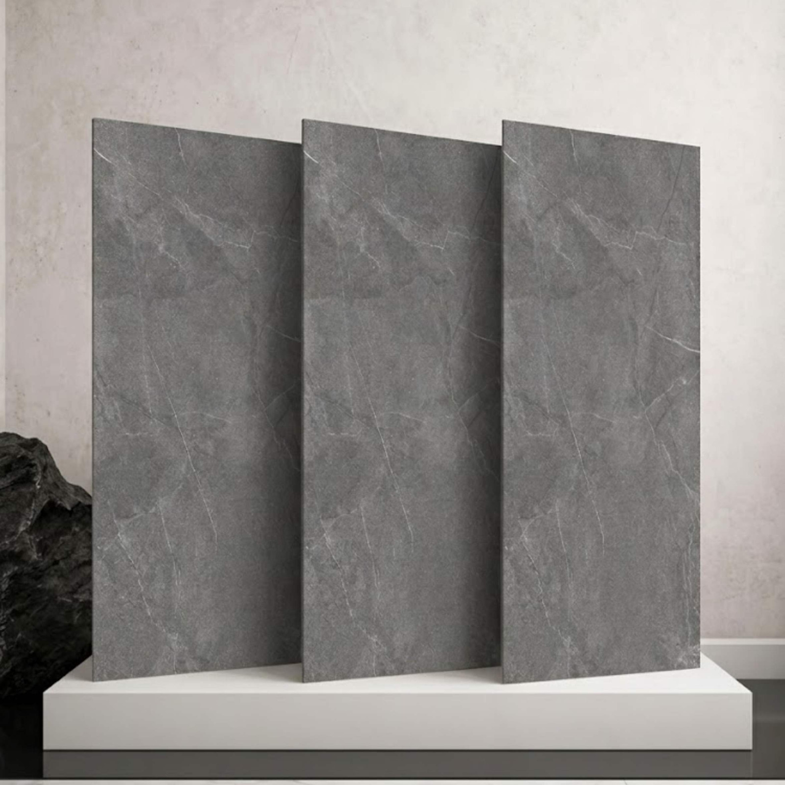 Three large rectangular grey marble-look tiles displayed upright side-by-side on a white block platform from alwakil.ae